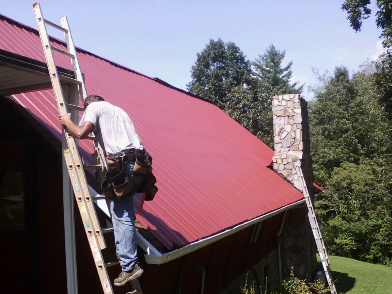Roofer on a ladder beside a red metal roof and stone chimney — Soffit & Fascia Repair by Roofing Brothers in Maple Shade